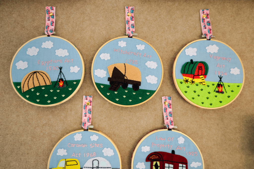 Circular embroidered hoops on a wall depicting historical anti-homelessness laws with colorful fabric illustrations of tents, caravans, and campfires.

