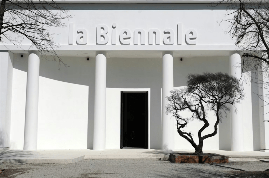 White architectural structures and signage for La Biennale di Venezia 2026 under clear skies, with trees and open pathways.

