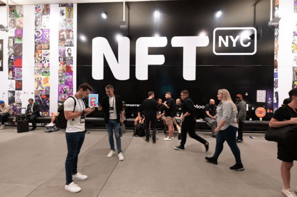 Busy indoor conference space at NFT.NYC with attendees walking and interacting in front of a large black wall featuring the white “NFT NYC” logo, surrounded by colorful NFT artwork displays.

