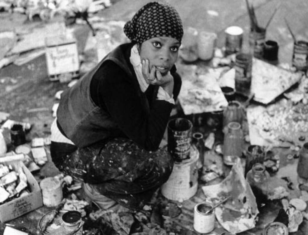 Black-and-white photograph of Mary Lovelace O’Neal seated in a cluttered artist’s studio, wearing a polka-dot head covering and looking directly at the camera with a thoughtful expression, surrounded by art supplies and canvases.