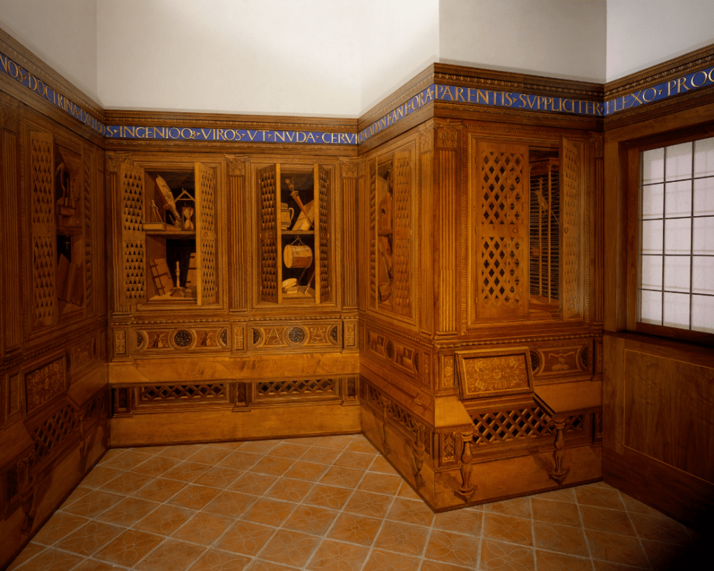  Renaissance wooden study room with trompe l'oeil intarsia panels depicting open cabinets, books, instruments, and symbolic objects in detailed wood inlay.

