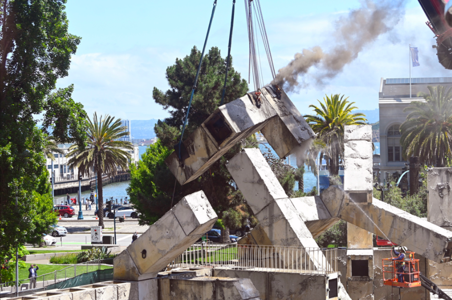 Crane lifting a massive concrete arm of the Vaillancourt Fountain with flames and heavy smoke rising during controversial dismantling in Embarcadero Plaza, San Francisco, May 2026.

