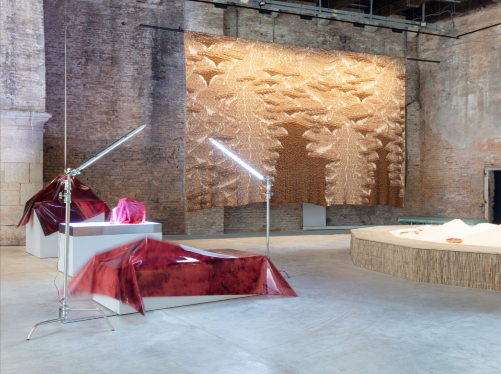 Interior art installation at Venice Biennale featuring illuminated red translucent sculptures, spotlights, and a large textured golden wall hanging in a historic brick venue.

