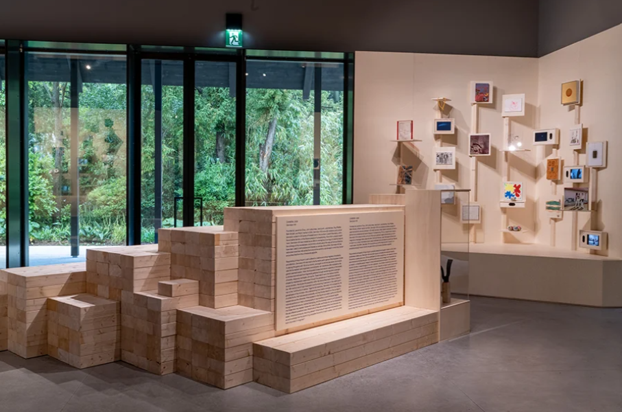 Light-filled gallery with stacked wooden block structures, large text panels, and small framed artworks arranged on walls.