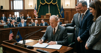 Governor sitting at desk signing document with flags and officials standing nearby