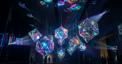 Hanging geometric prisms glowing with refracted colorful light beams in a dark room with viewers in the background