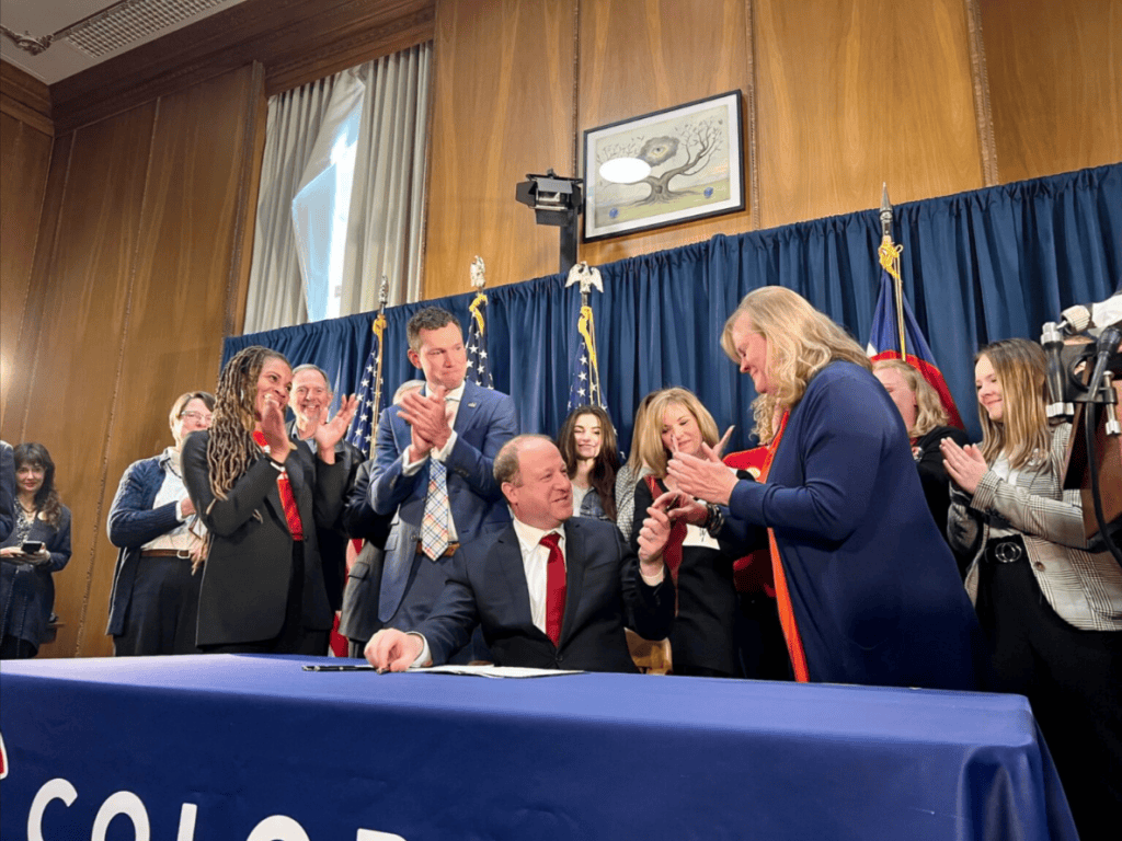 Colorado Governor Jared Polis at a bill signing ceremony with diverse group of attendees applauding)