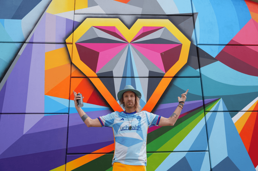 Man in hat holding spray can posing confidently in front of large geometric heart mural in urban setting)