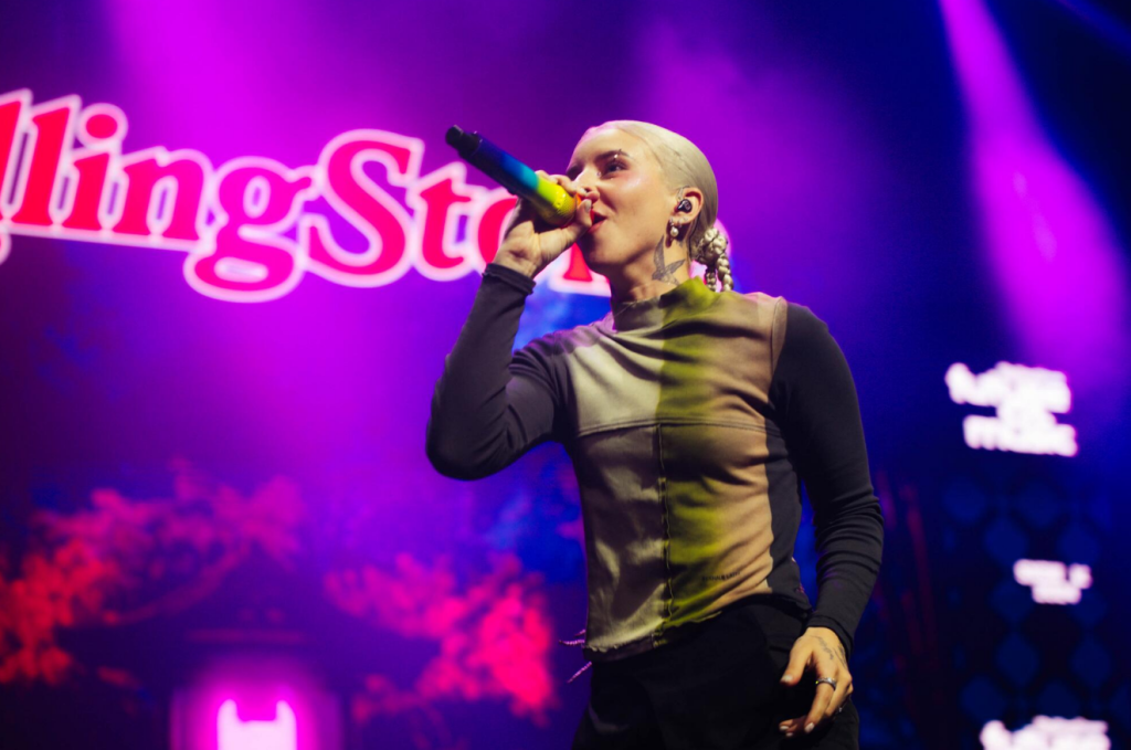 Young Miko on stage singing into a colorful microphone, showcasing neck and arm tattoos against purple concert backdrop.