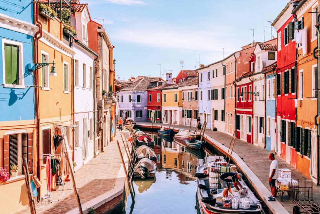 Vibrant colorful buildings lining a canal in Venice during peak summer heat with boats and pedestrians visible.