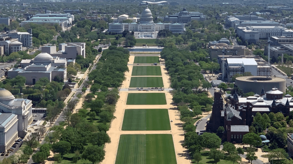 Expansive aerial photograph showing the green expanses, reflecting pool, Capitol Building, and surrounding federal architecture of the National Mall under clear skies.