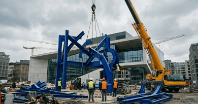 Workers assembling a large blue sculpture with a crane outside an arts center
